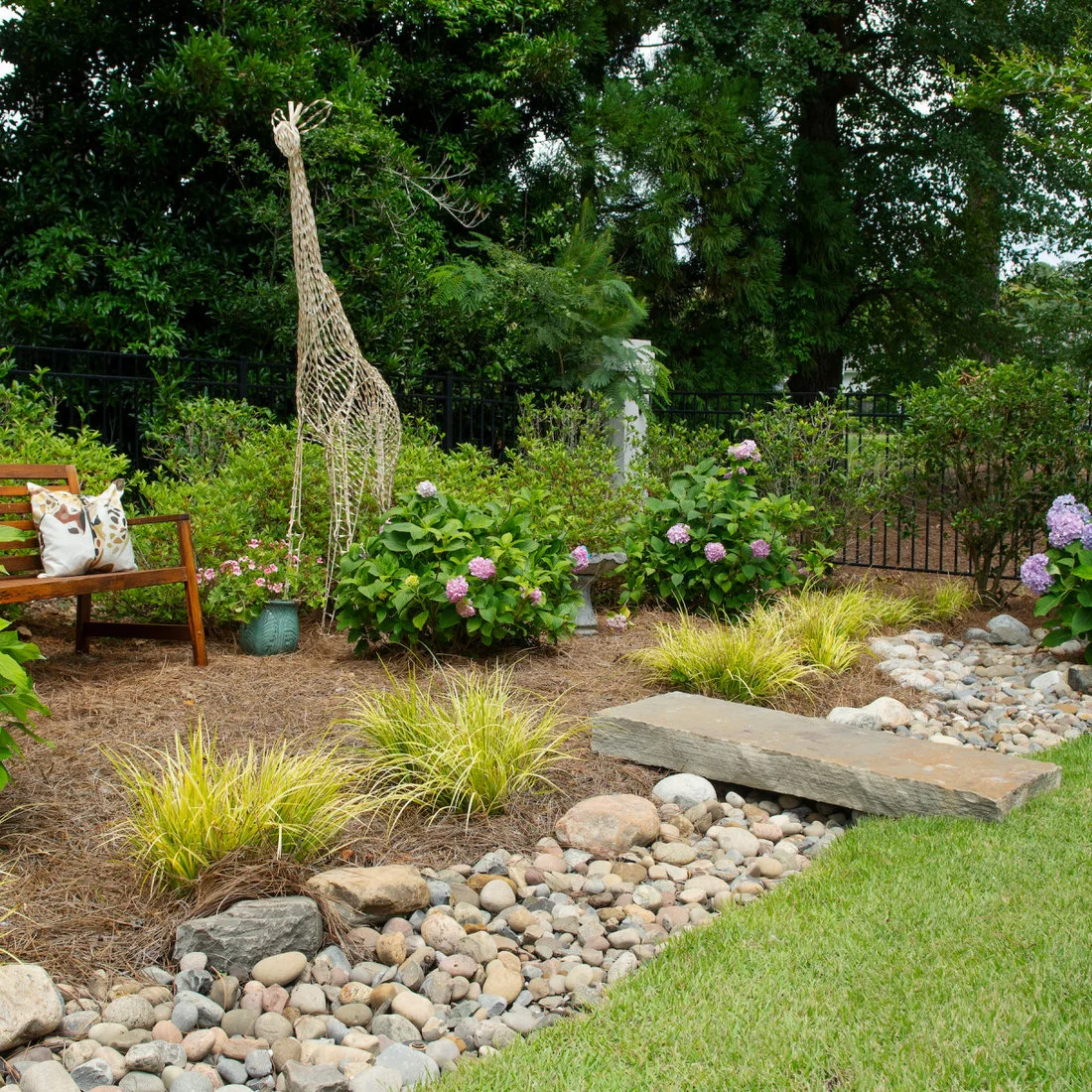A flat stone bridge crosses a small river-rock channel beside grasses and a garden bench