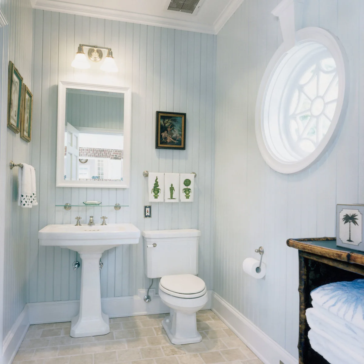 A white pedestal sink stands against pale blue vertical paneling, with a narrow glass shelf below a rectangular mirror and a round side window.