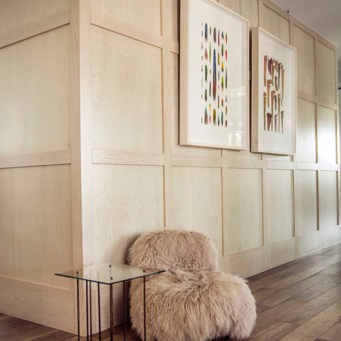 Pale wood wall panels and framed art line a hallway beside a small shaggy chair and glass-topped side table.