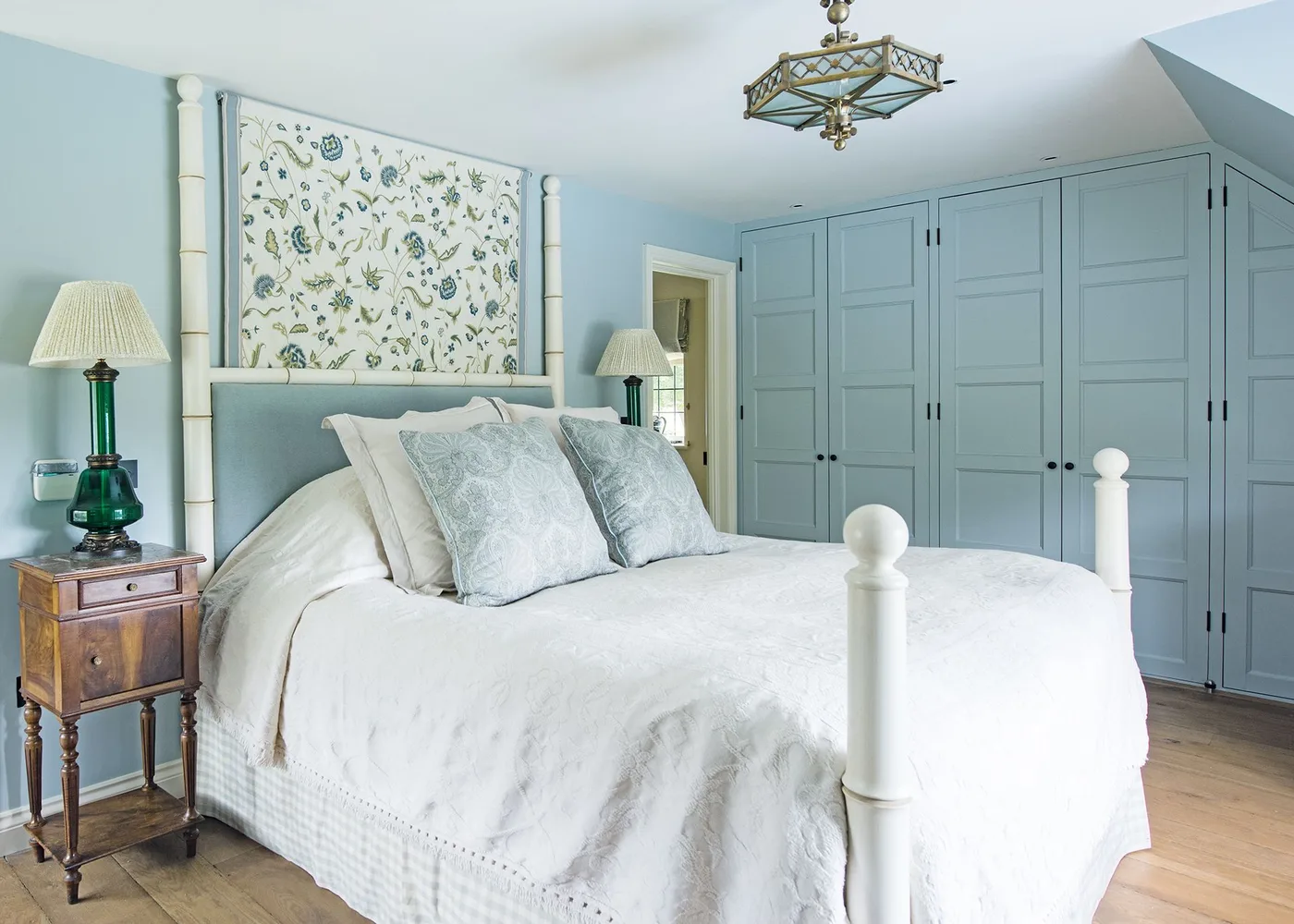 Pale blue bedroom with paneled fitted wardrobes, a white post bed, patterned headboard insert and wooden bedside tables.