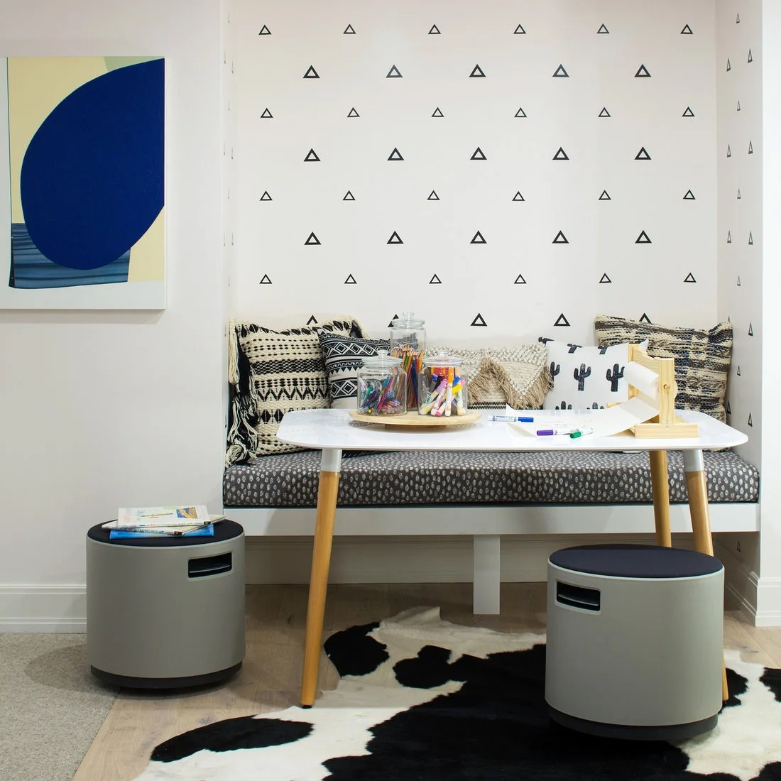 A white table with wood legs stands before a patterned built-in bench, with pencil jars, a paper roll, gray storage stools and small triangle wall motifs.