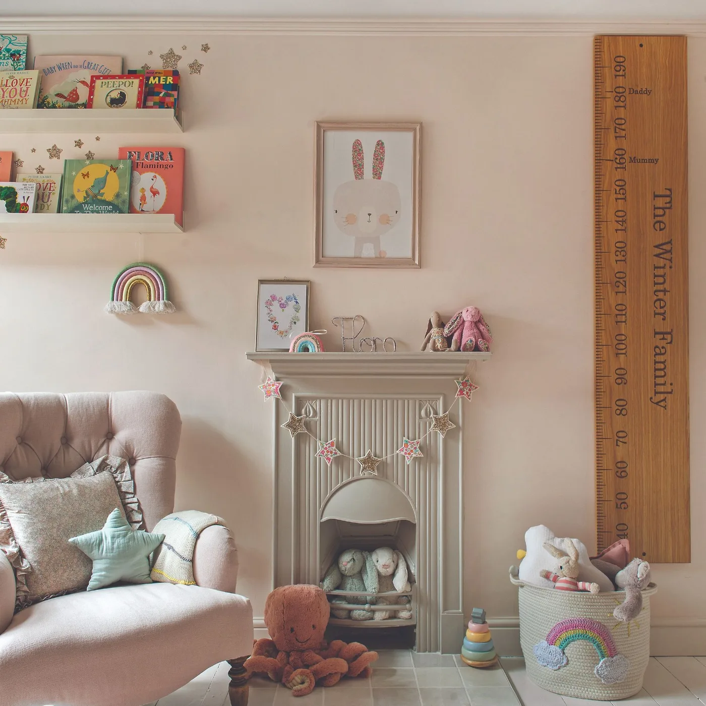 Pale pink upholstered chair beside cream fireplace, framed animal pictures, books and tall wooden measuring piece.