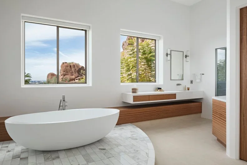 Low timber bathroom storage sits beneath white counters beside a pale tub and windows overlooking rock and trees.