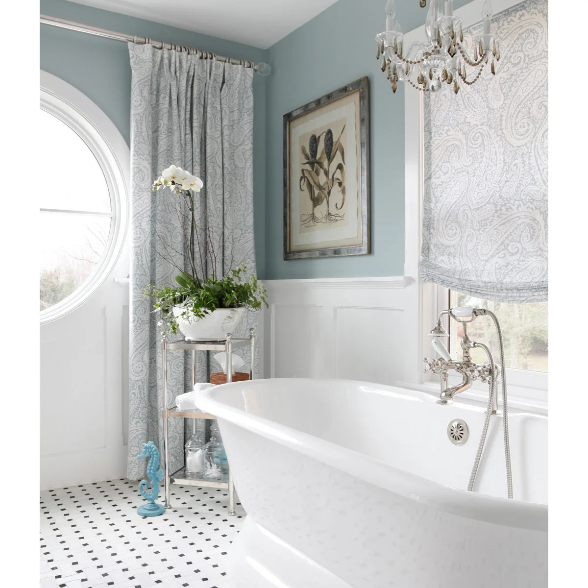 Pale patterned drapes frame a round window beside a matching Roman shade above a white freestanding bath.