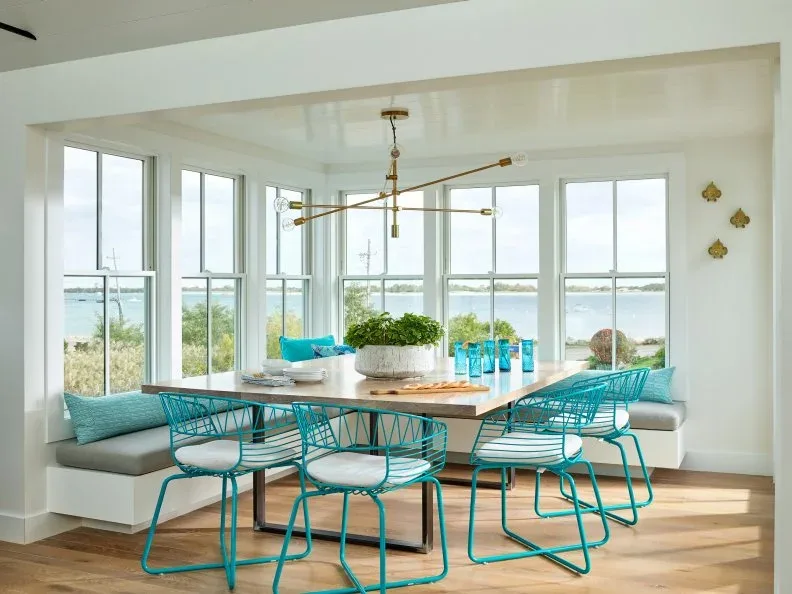 Dining nook with turquoise open-frame chairs, white wall bench, wooden table and wide windows overlooking water.