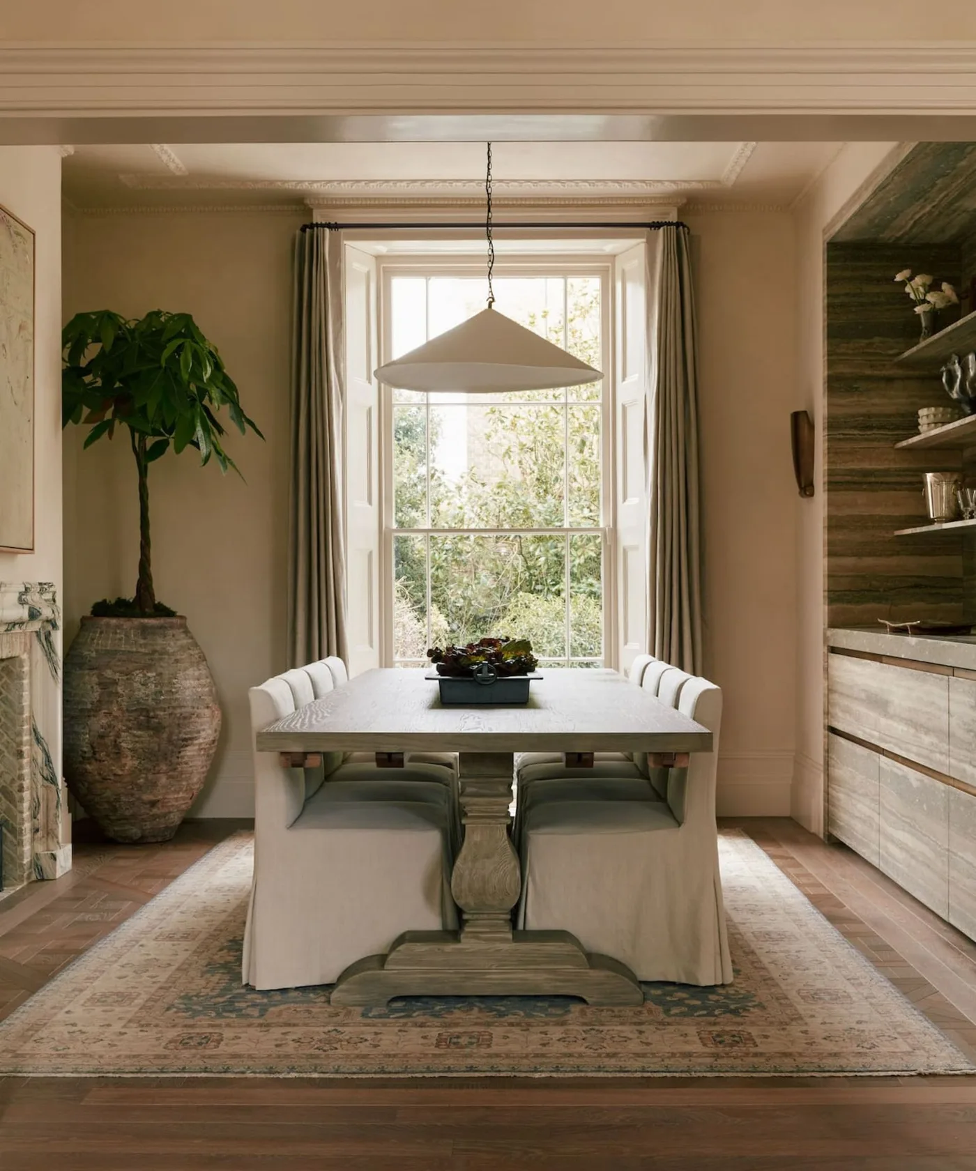 Country dining room with pale skirted chairs around a wooden pedestal table, recessed wooden storage and a plant in a large textured container.