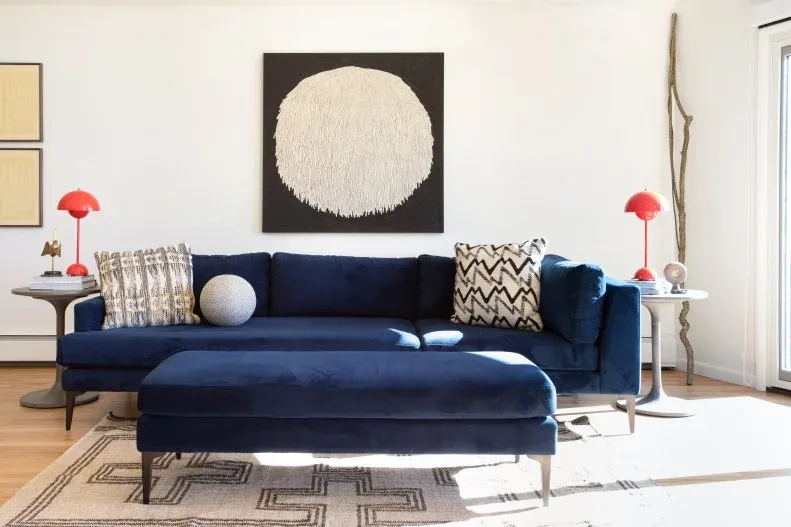 Living room with deep blue sofa and matching ottoman, red dome-shaped lamps, pale side tables, patterned cushions and large circular abstract artwork.