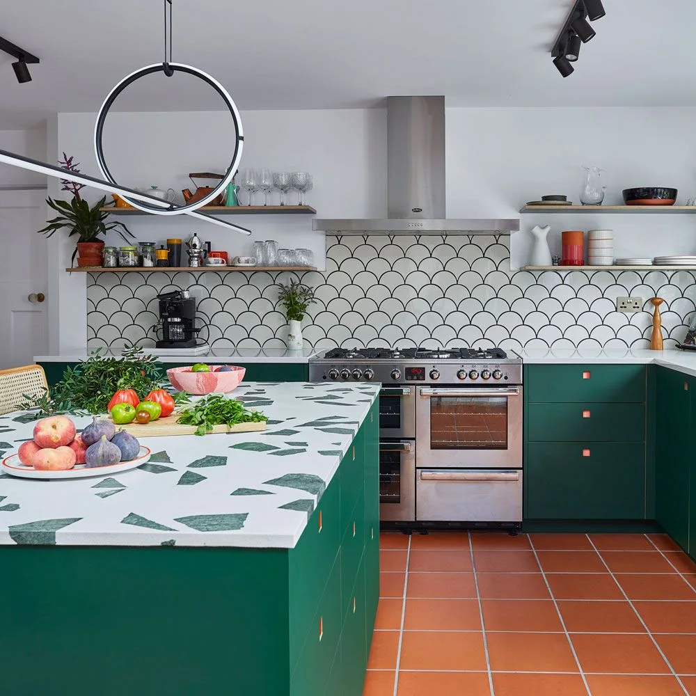 Green flat-fronted cabinets sit below a pale counter with green fragments beside scalloped wall tiles and open wooden shelves