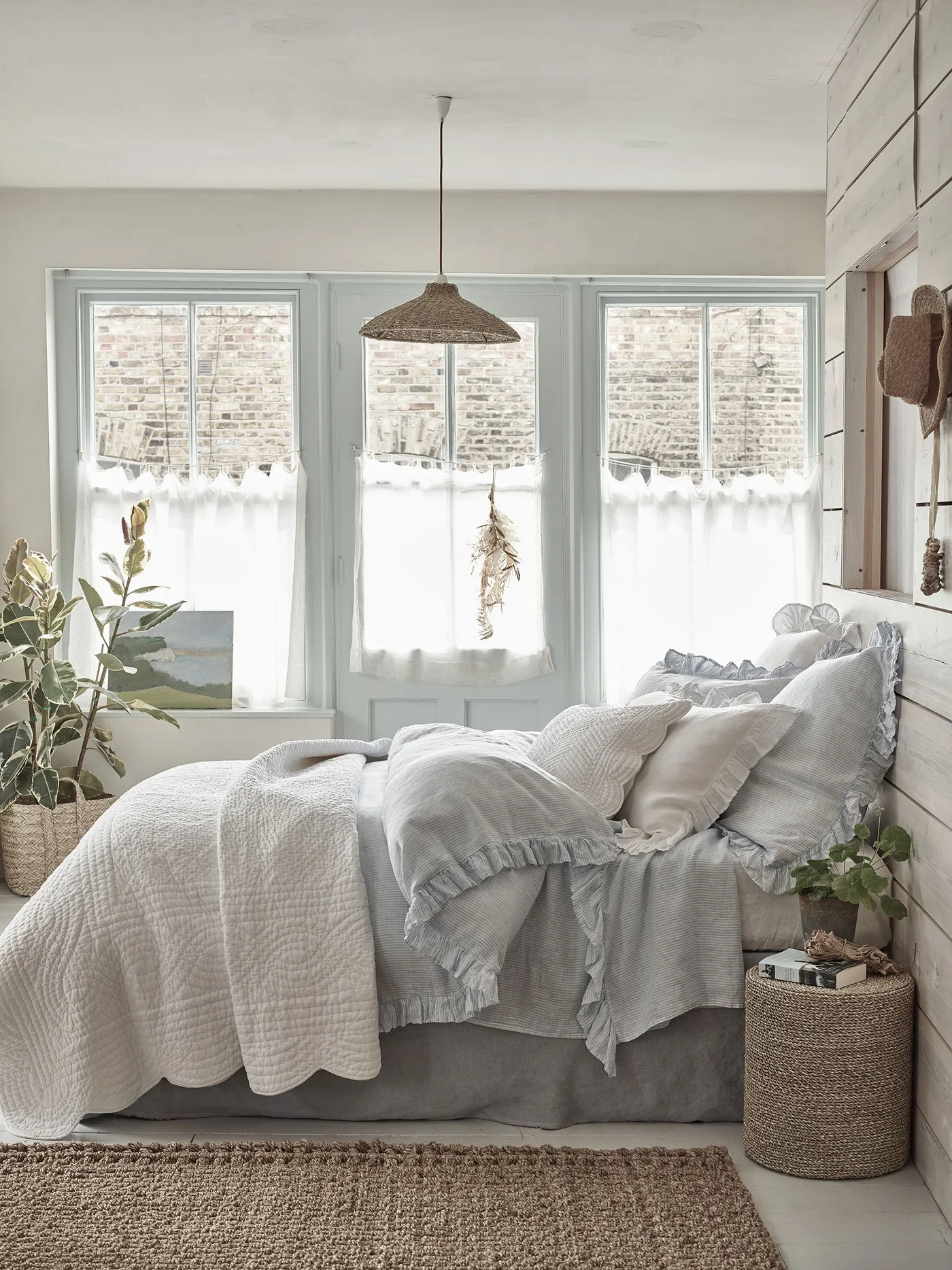 Pale country bedroom with layered white and light gray bedding, lower-window curtains, a woven pendant shade and a small round woven bedside surface.