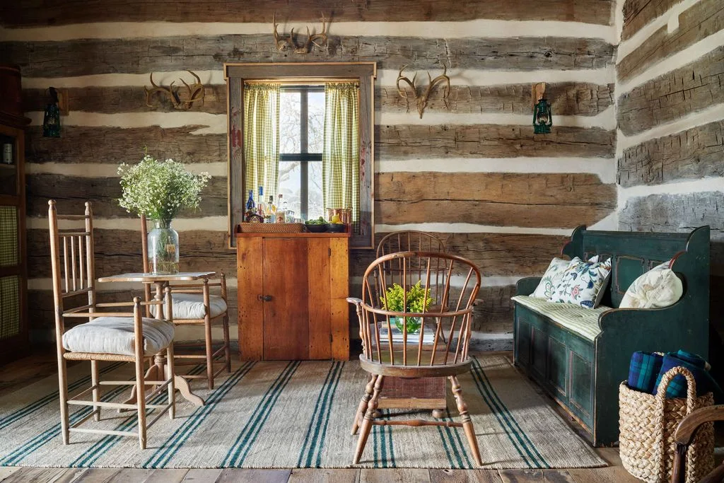 Log-walled sitting area with a dark green bench, wooden chairs and a striped rug
