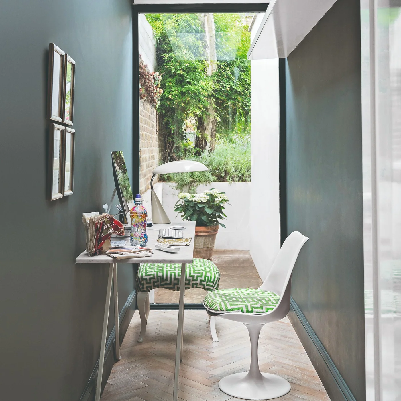 A slim desk and white chair with green patterned upholstery stand in a narrow blue-gray space opening toward greenery and overhead glazing.