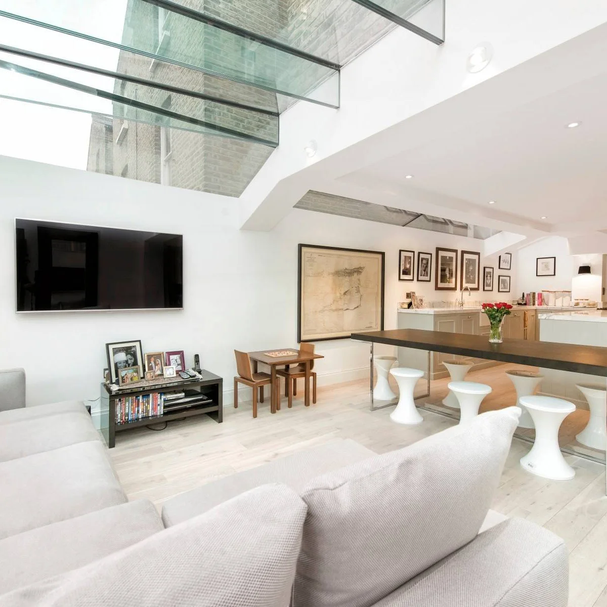 A pale sofa faces a wall television beneath roof glazing, with a long dark dining table and white stools beside the kitchen.