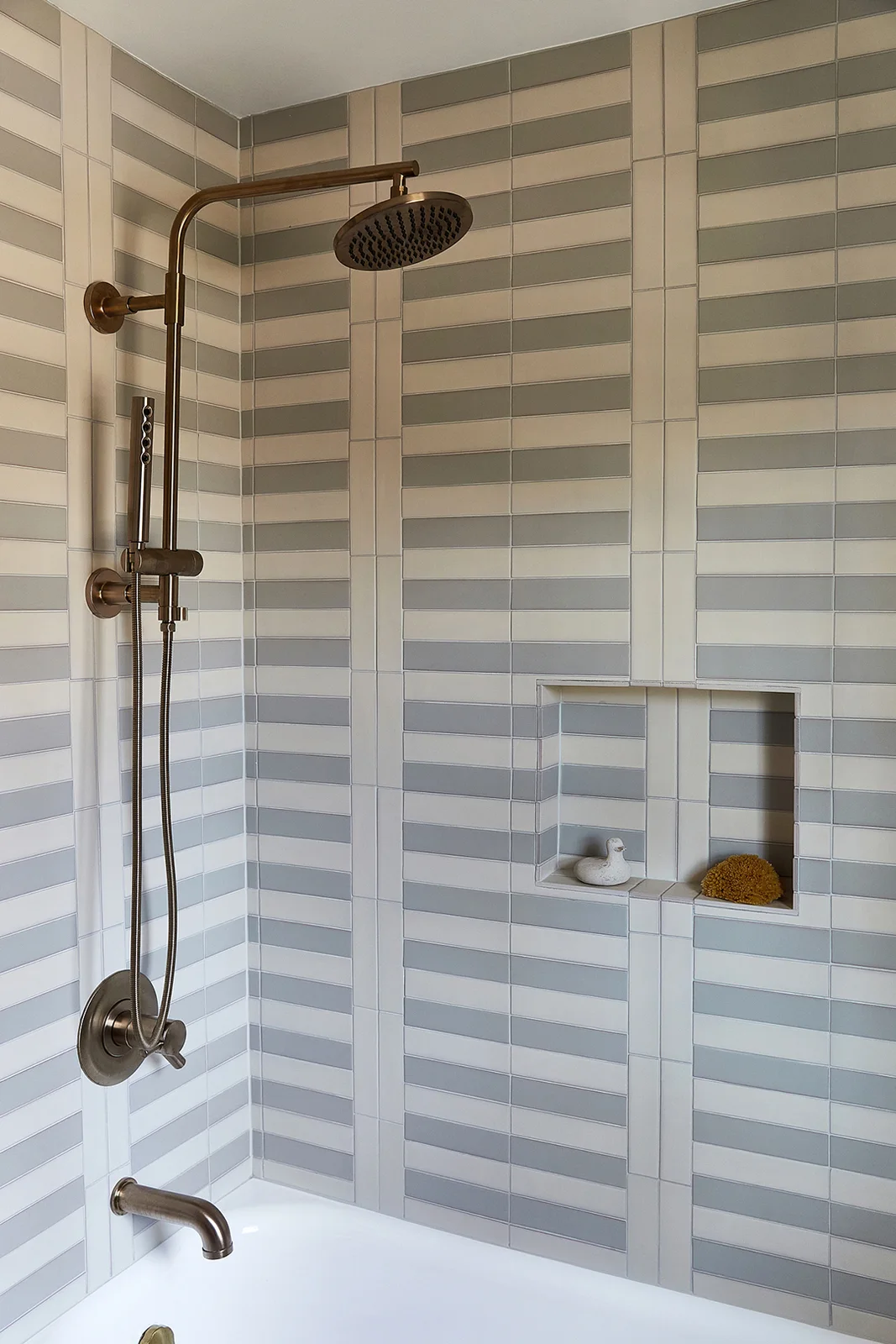 Gray and pale rectangular tiles form horizontal shower stripes around brass-colored fittings and a recessed shelf