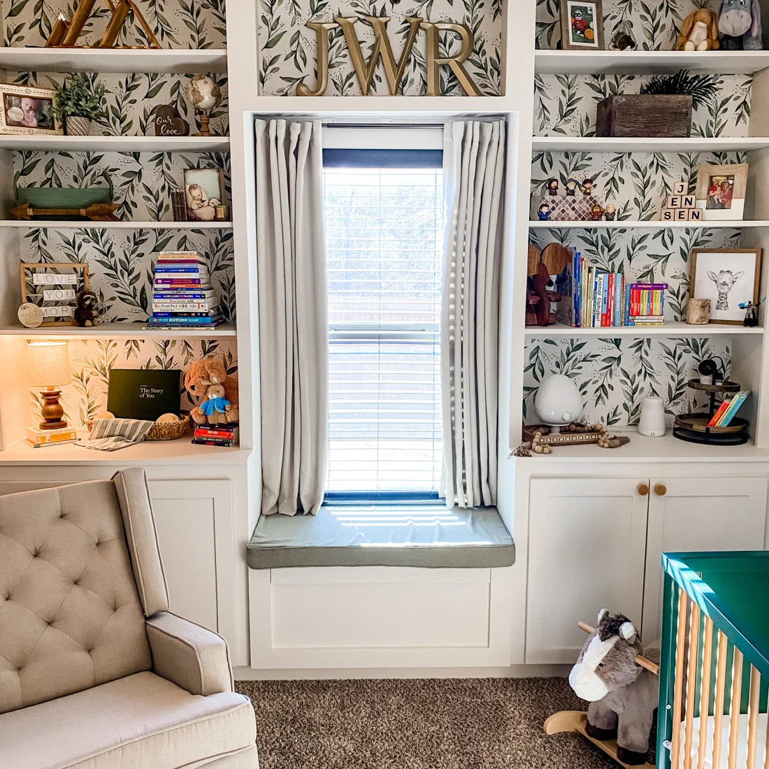 White shelves with leafy backing flank a narrow window seat, with books, toys and framed pictures arranged around a gray chair.