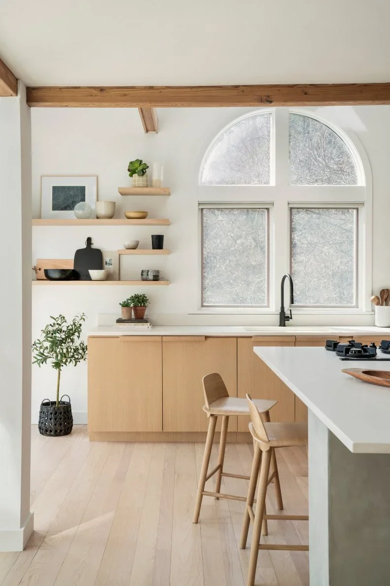 Pale wood kitchen cabinets and small floating shelves beside an arched window and white island