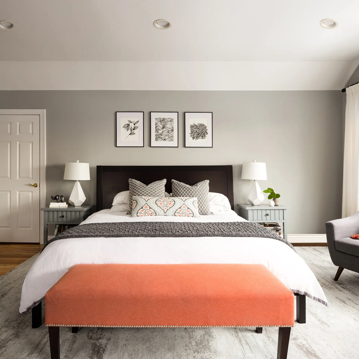 Three small black-framed botanical and patterned prints above a dark headboard with white lamps and an orange bench.