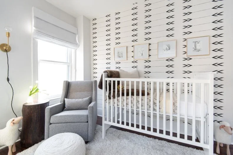 A white nursery crib and gray armchair stand beside a pale wall with repeated dark arrow shapes, a small wood table and a white rounded footrest