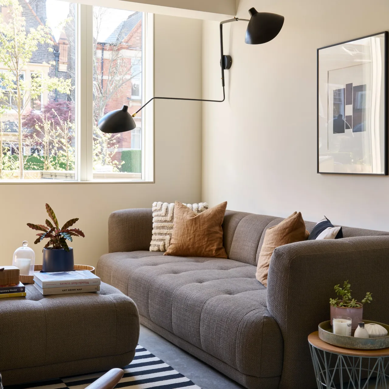 Warm gray sofa and matching ottoman in a warm cream room with rust cushions, a large window and dark wall lights