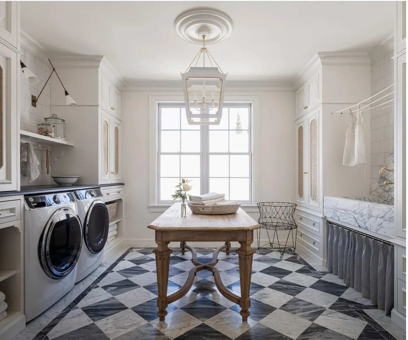 Pale laundry room with central turned-leg wood table, side-by-side machines, checked floor, lantern pendant and skirted sink