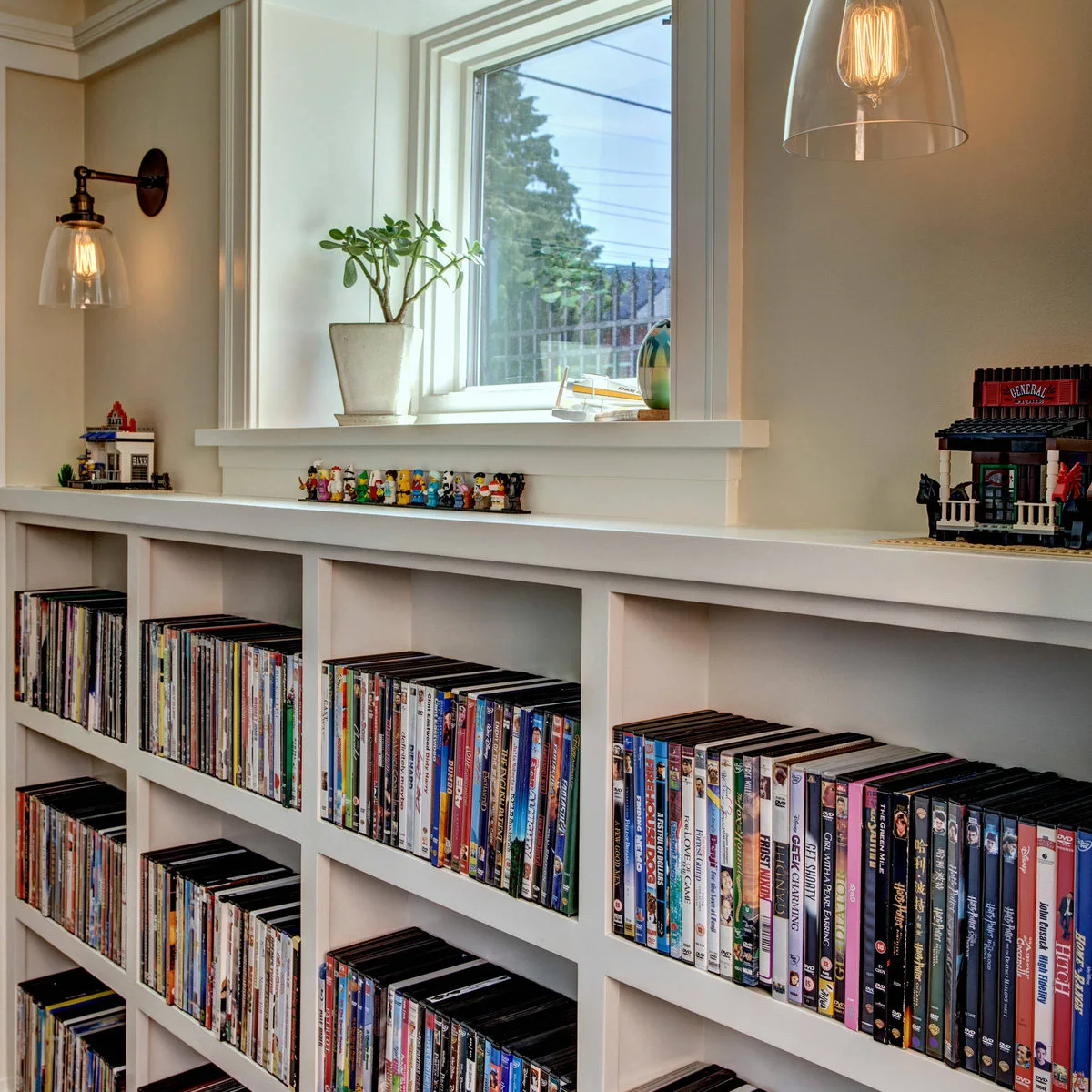 A dark short-arm wall light with a clear bell-shaped shade sits above a white bookcase filled with books and small models.