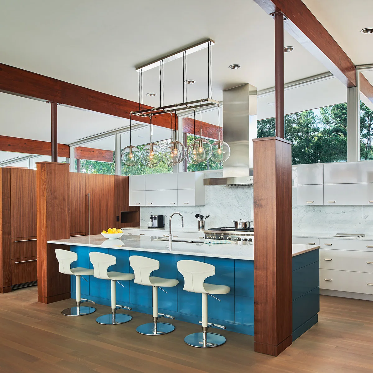 A vivid blue island with white swivel stools in a kitchen with wood beams, tall wood cabinets and high windows.