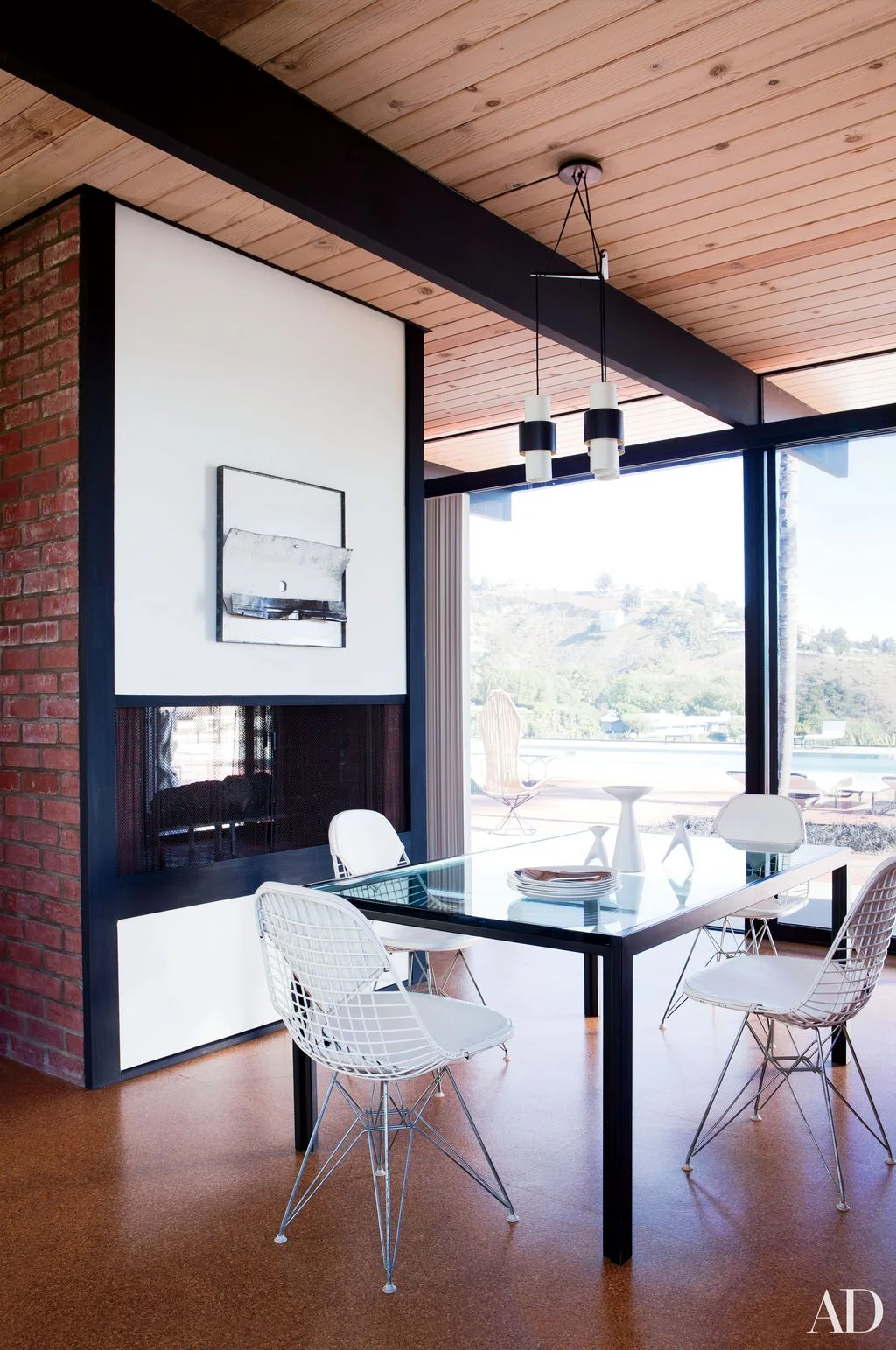 A dark fireplace opening sits below a plain white wall panel and framed artwork beside a brick wall, wood ceiling, glass dining table and white wire chairs