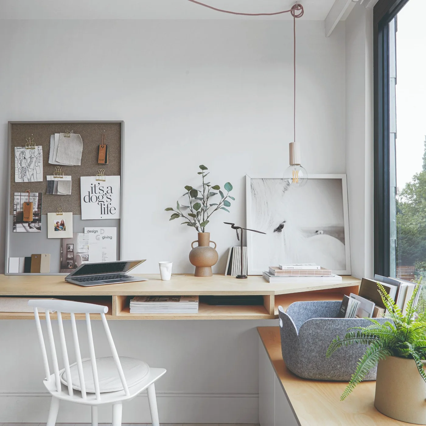 A pale wood corner workspace has open slots beneath the desktop, a white spindle-back chair, a noticeboard, a dark-framed window and a gray felt file basket.