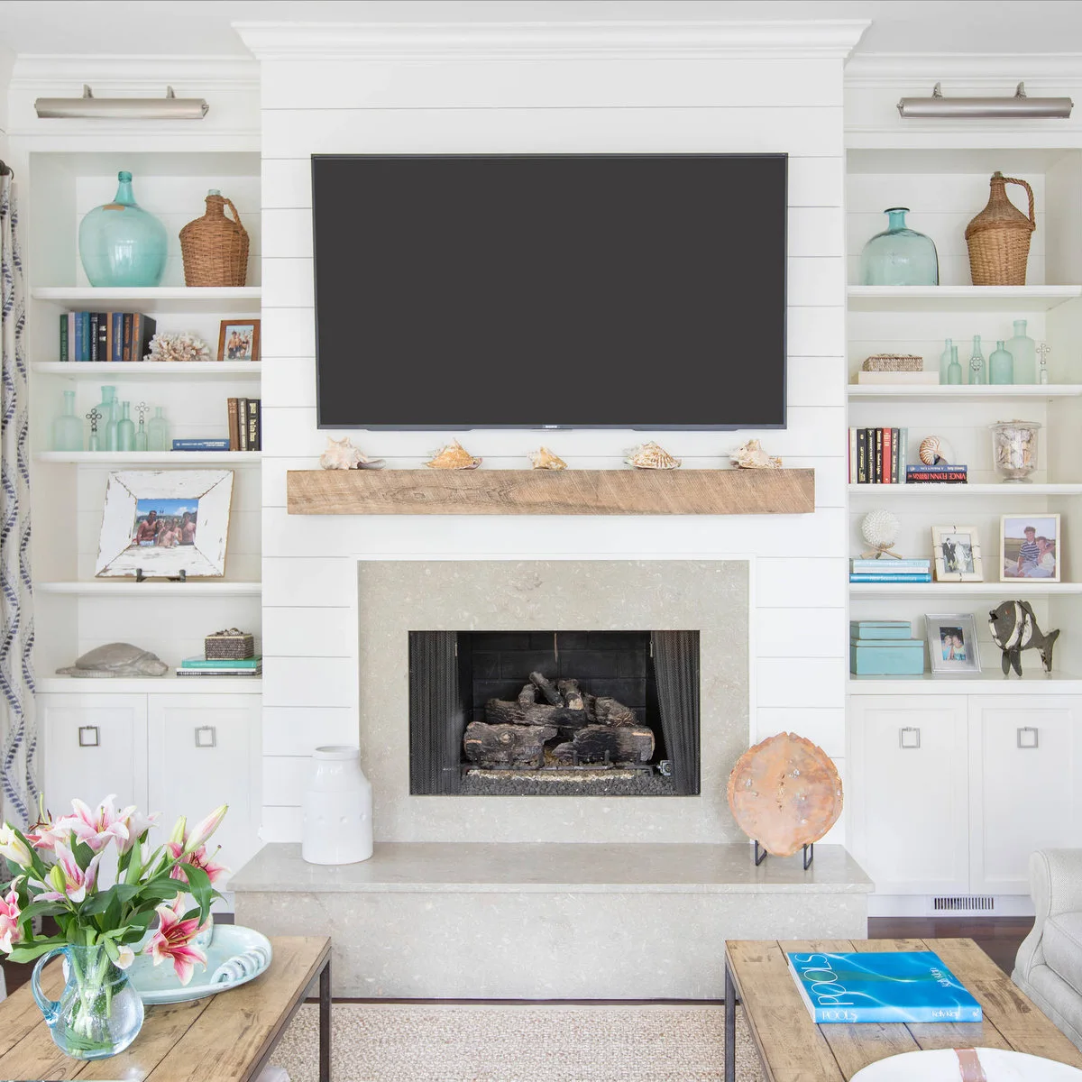 A television above a wood mantel and pale fireplace, with white shelves displaying blue-green bottles, books and woven vessels.