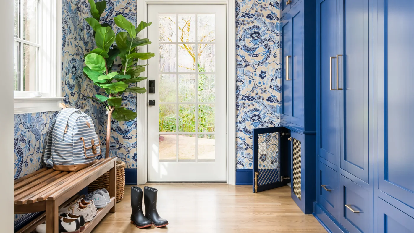 Tall blue mudroom cupboards and lower drawers stand opposite a slatted wood bench beside blue patterned wallpaper and a glass door.