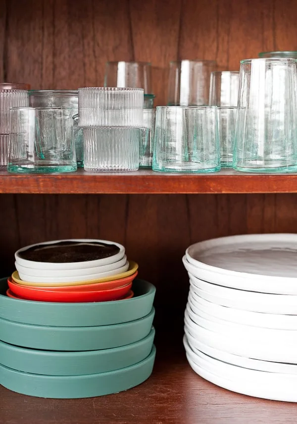 An open wood sideboard compartment holds drinking glasses above separate stacks of pale and green dishes on a lower shelf