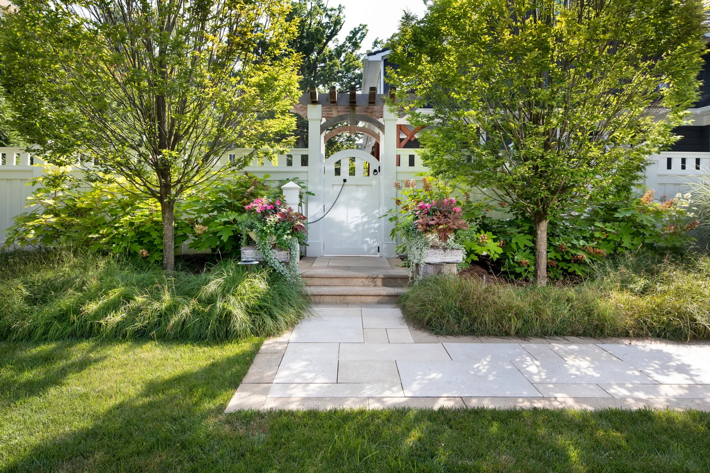 Pale paving widens at steps beneath a white garden gate framed by trees and planted pots