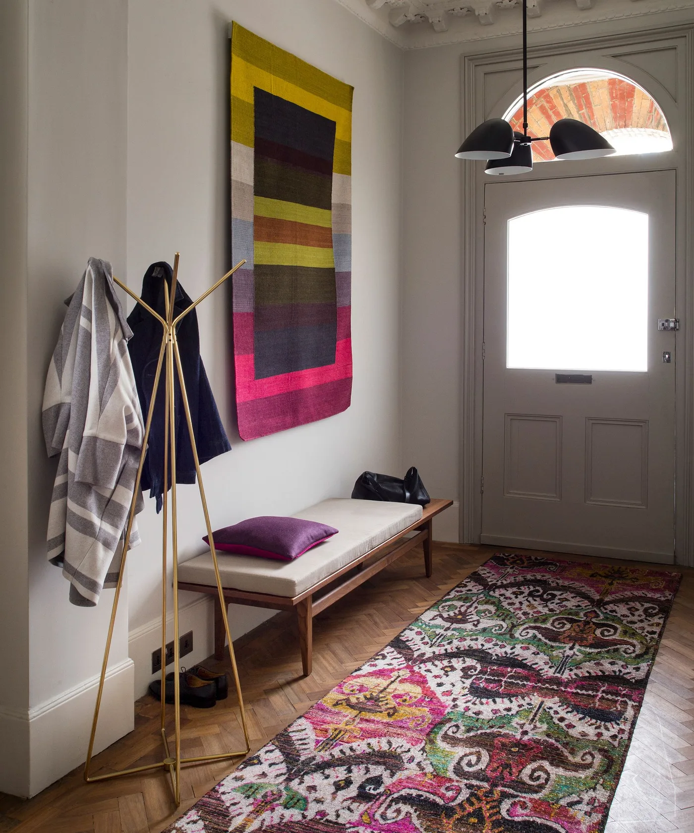 Colorful patterned runner beside a long pale-cushioned wood bench, gold-toned coat stand and glazed gray front door