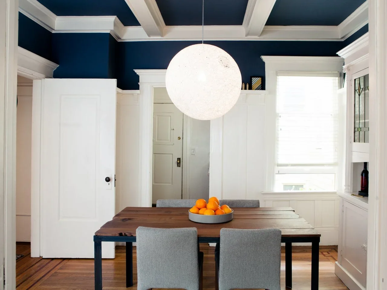 A dining room has navy upper walls and ceiling panels, white beams and wainscoting, a large globe pendant and a timber table.