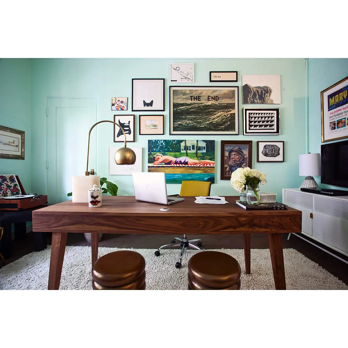 Broad wood desk with yellow chair, two visitor stools and turquoise walls covered in framed art
