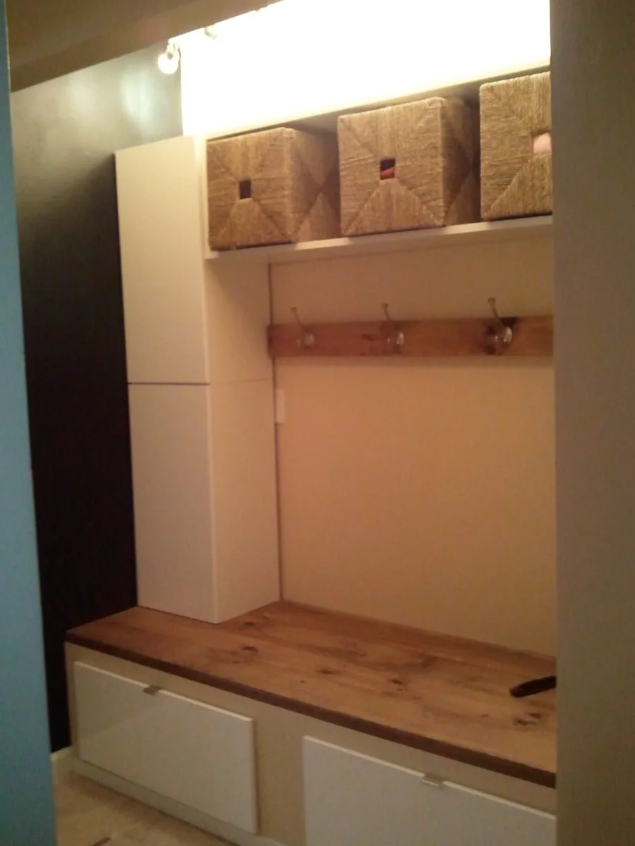Compact white mudroom storage with three upper woven baskets, a wood hook rail, wood seat and two lower drawers.