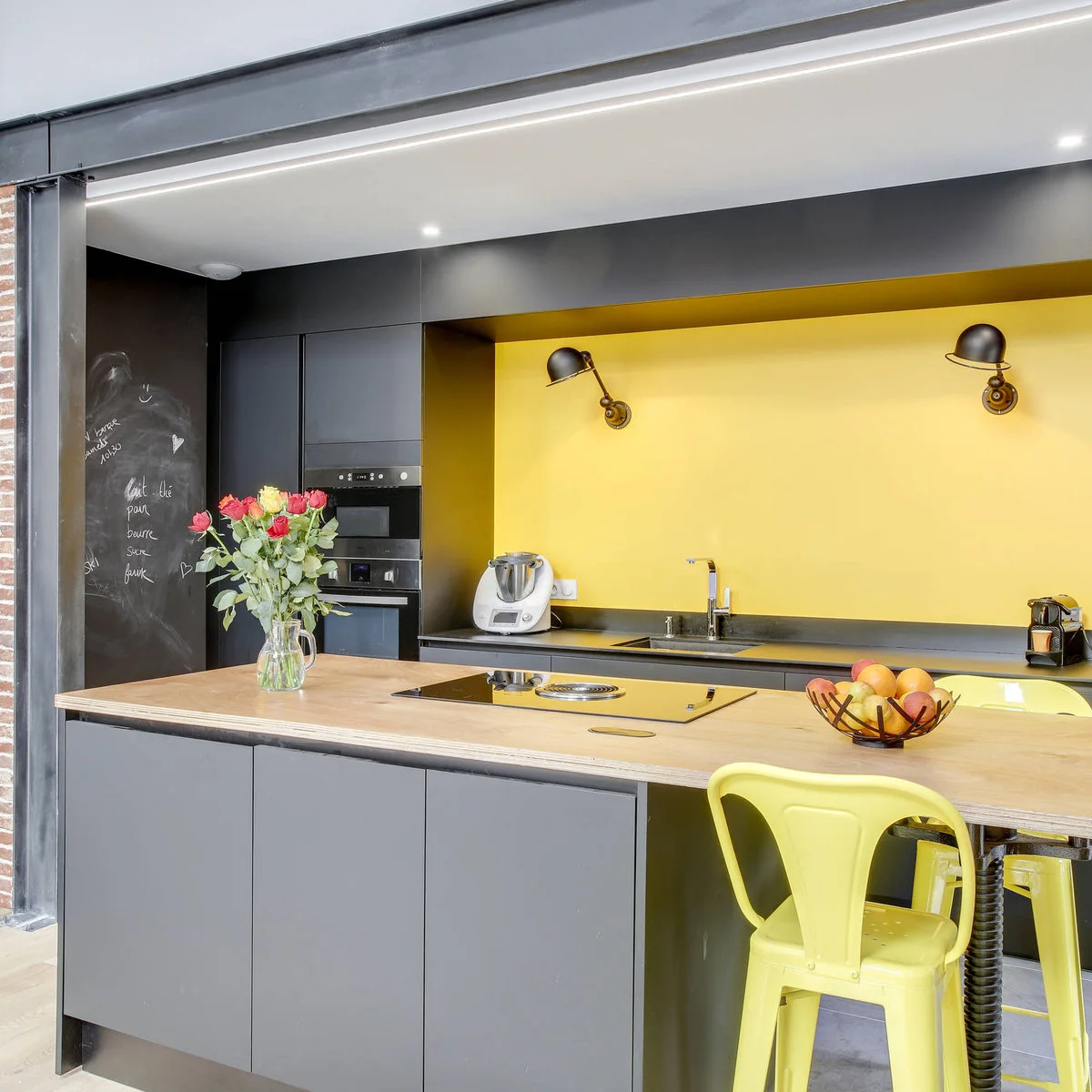 Dark gray kitchen cabinets around a yellow recessed wall with black lamps and yellow stools