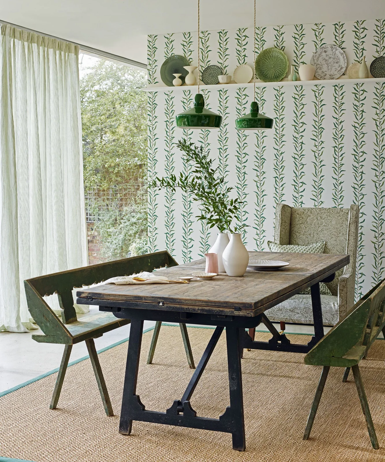 Leaf-patterned wallpaper sits behind a wooden table and benches beneath green pendant lights and a shelf of decorative plates