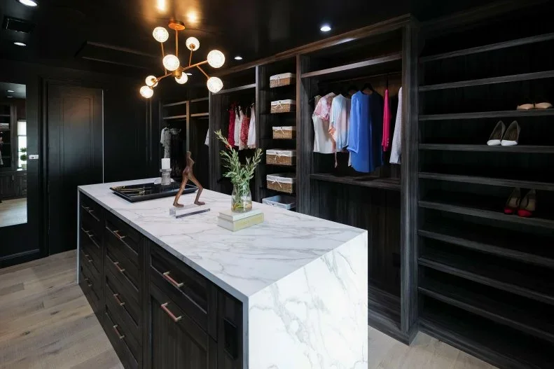 Dark open closet cabinets surround a drawer island with a pale veined top and a branching globe light.