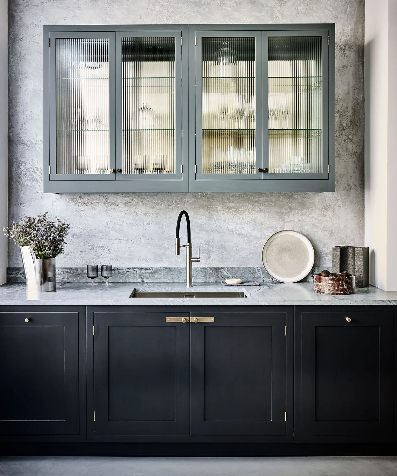 Dark lower cabinet fronts sit beneath a pale veined counter and sink with a gray framed textured-glass wall cupboard and dark faucet