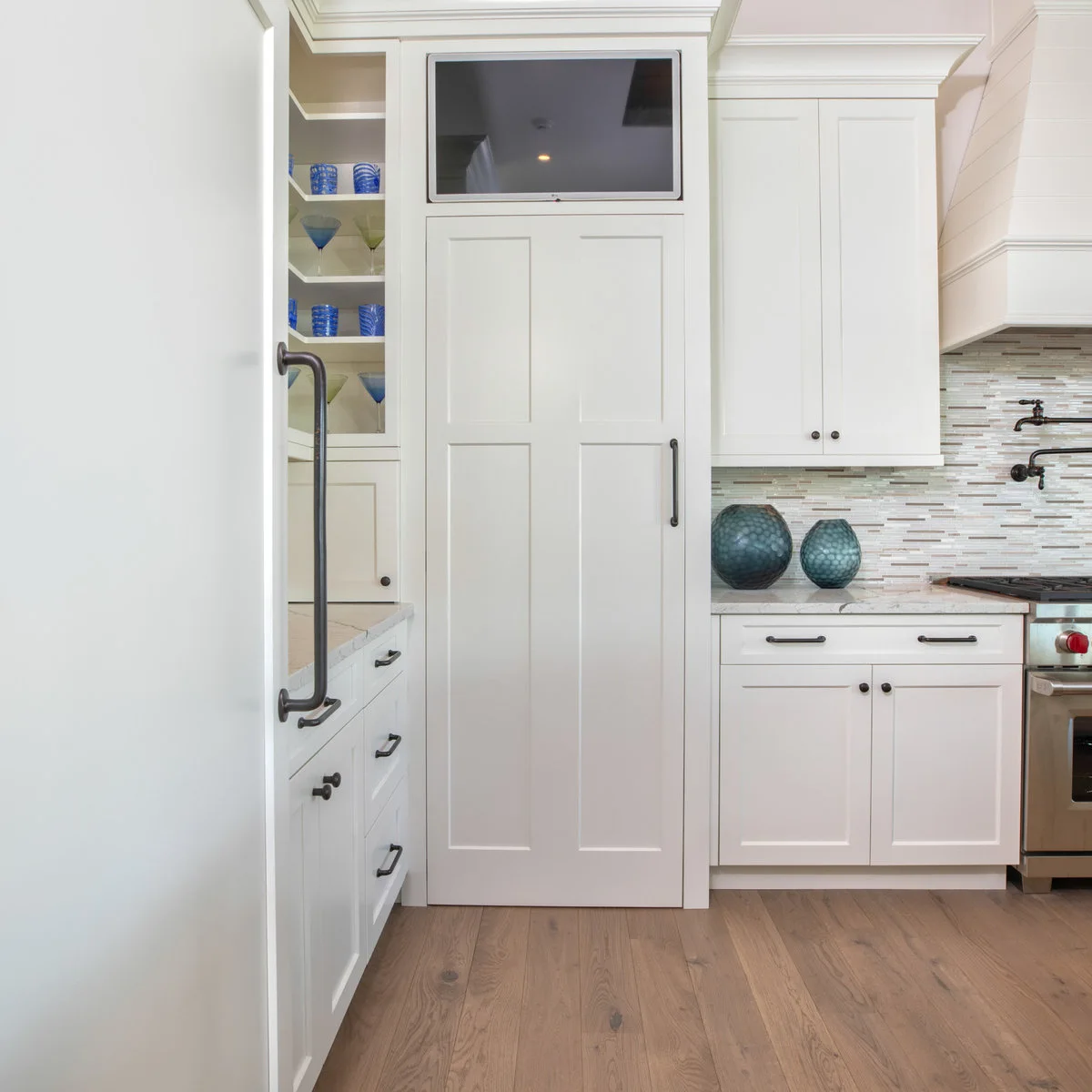 A tall white paneled pantry door with a dark handle sits between display shelves and a kitchen counter beneath a small screen.