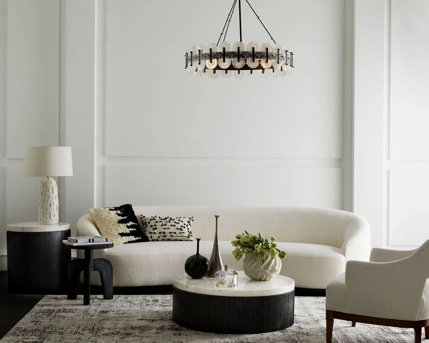 Pale living room with a curved white sofa, white armchair, dark round coffee table and a dark chandelier.