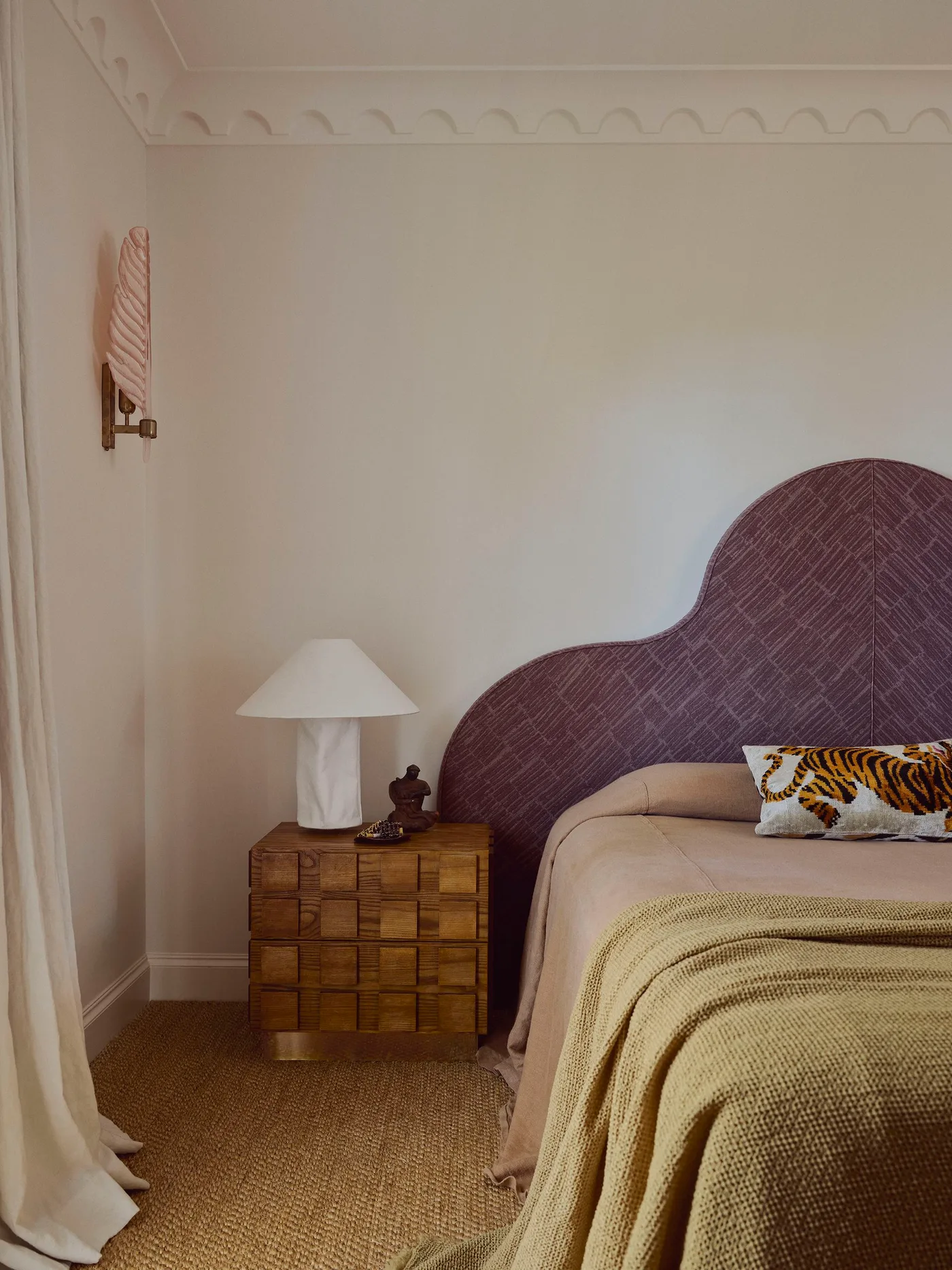 Warm pale bedroom with a curved aubergine-patterned headboard, tan and pink bedding, a khaki blanket and a block-patterned wood bedside cabinet