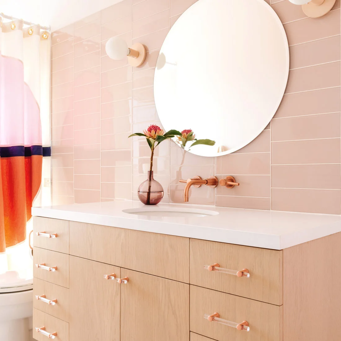 Pale pink rectangular tiles sit behind a round mirror and white sink above light wooden drawers