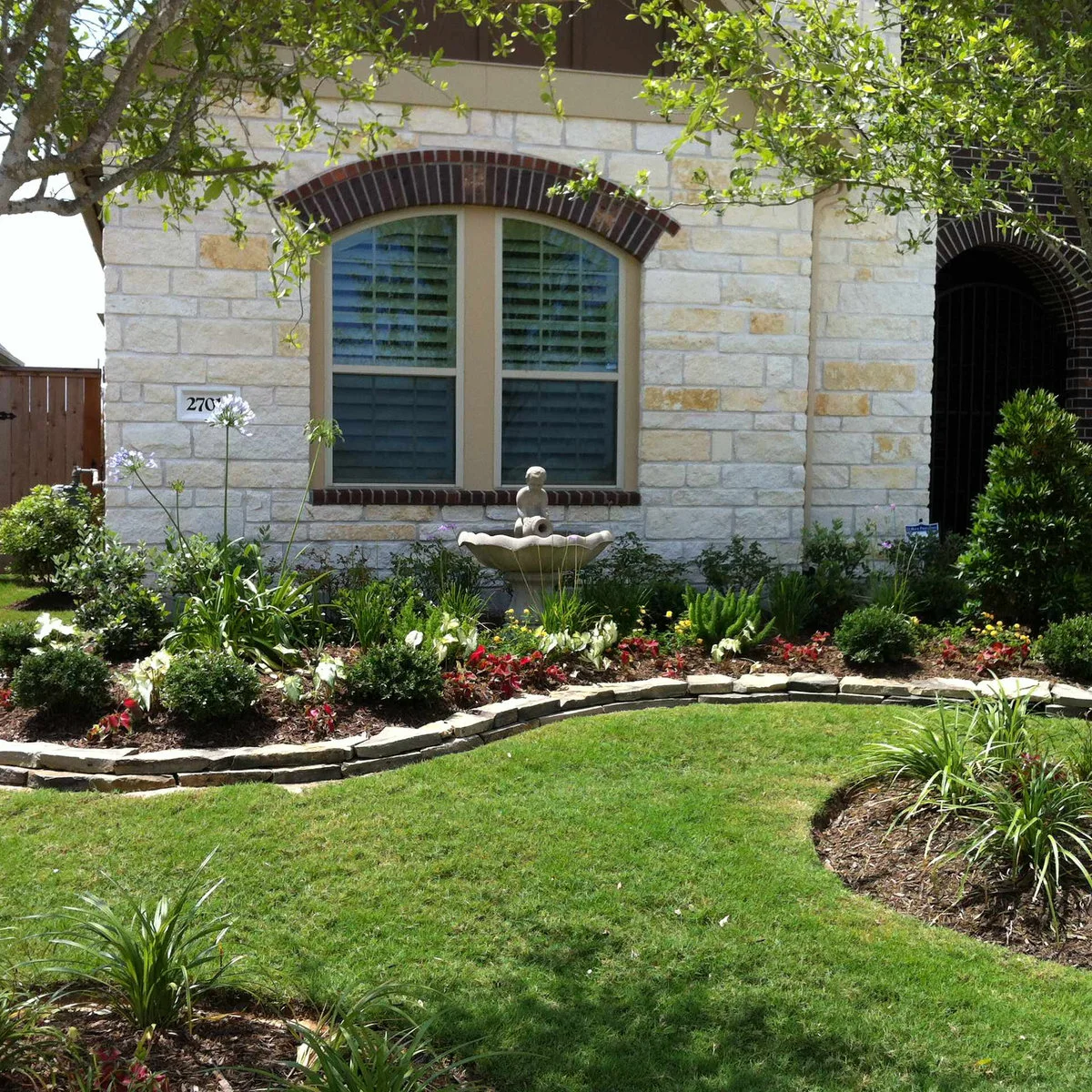 A small fountain beneath an arched front window, surrounded by low shrubs, colorful bedding and a curved lawn edge.