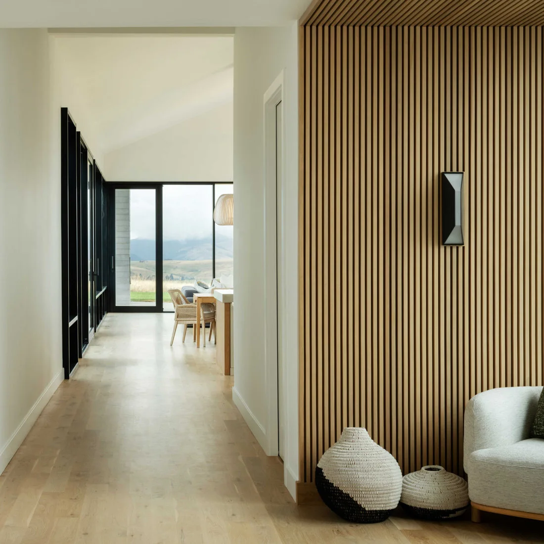 A wood slat wall with a black wall light borders a pale hallway leading toward dark-framed glazing and a dining area.