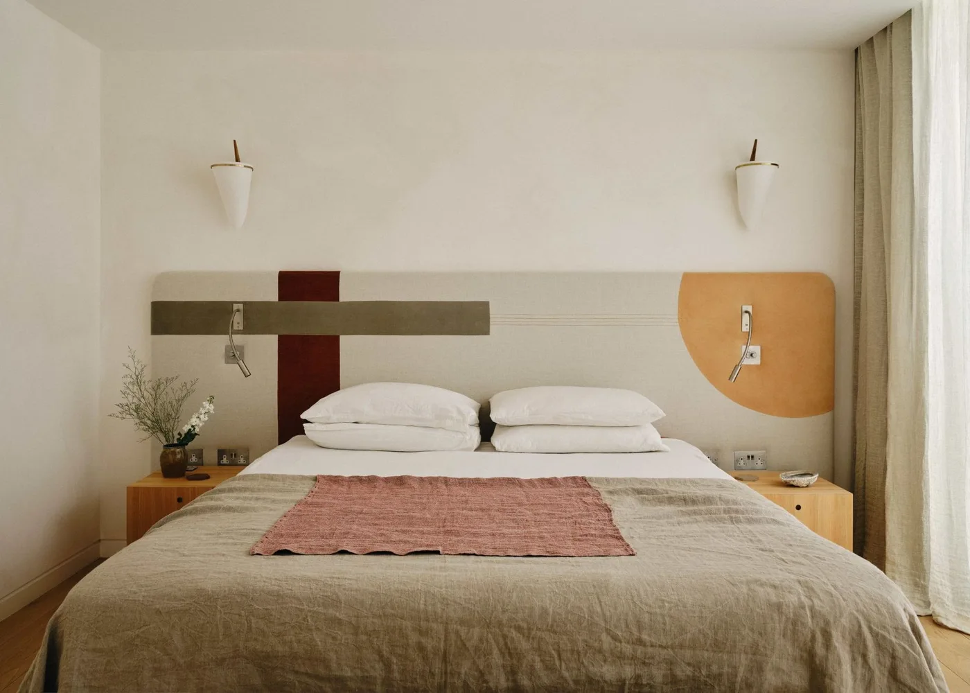 Bedroom with a long headboard patterned in olive, deep red and mustard, pale wood bedside shelves and white wall lights