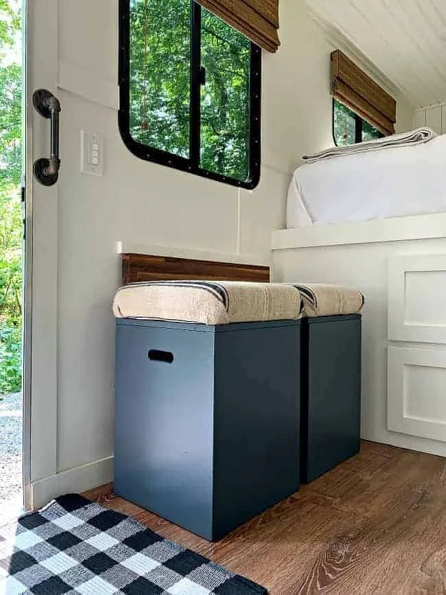Two blue storage boxes with upholstered lids sit beneath a camper window beside the entry door.