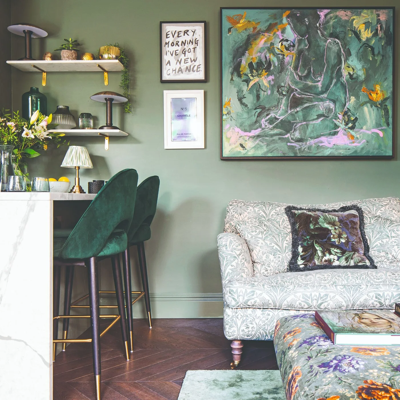 A patterned pale sofa sits beside green counter stools beneath artwork and shelves on a muted green wall.