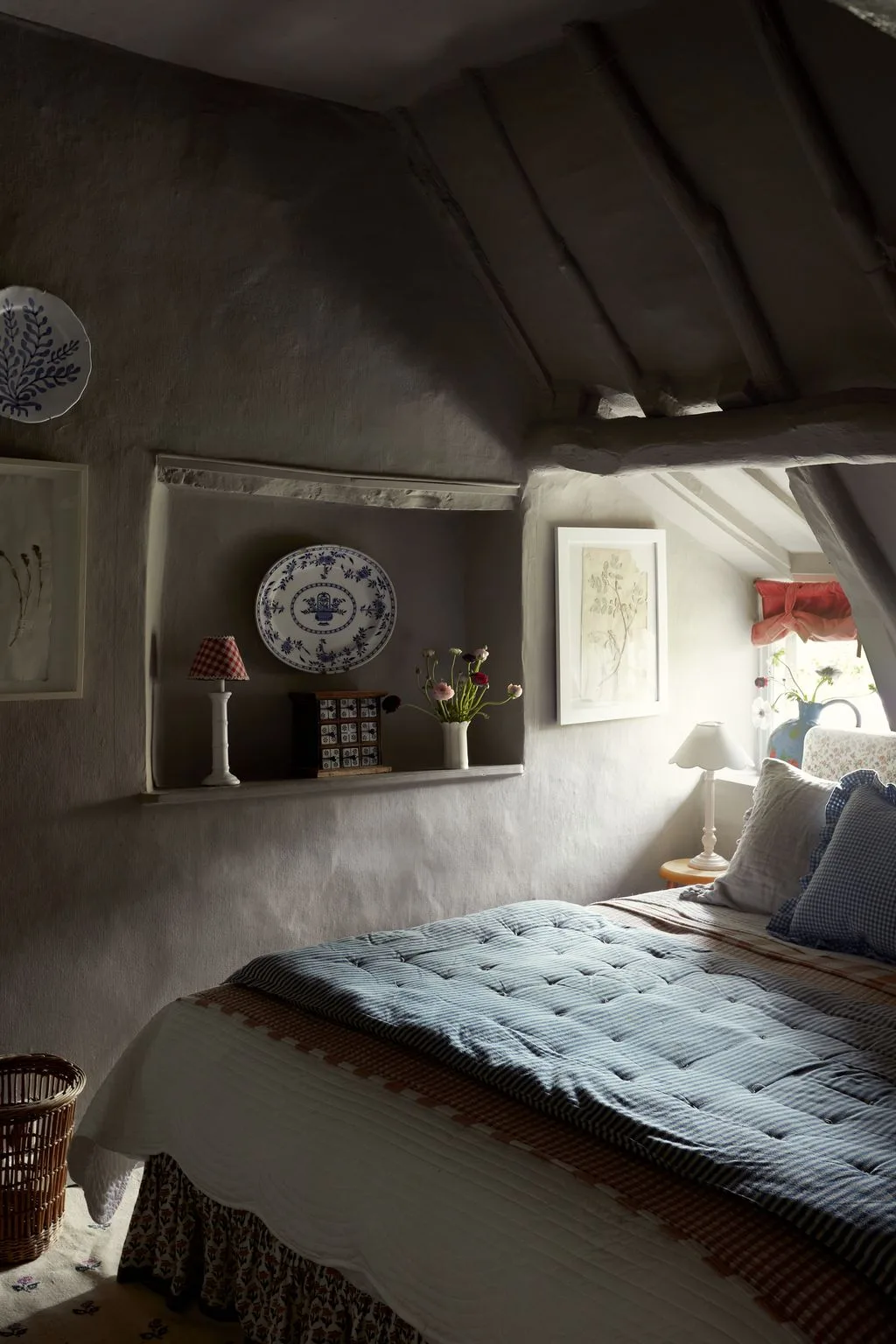 Dark attic bedroom with muted walls and roof beams, recessed display shelf, blue striped padded cover, small window and pale bedside lamp