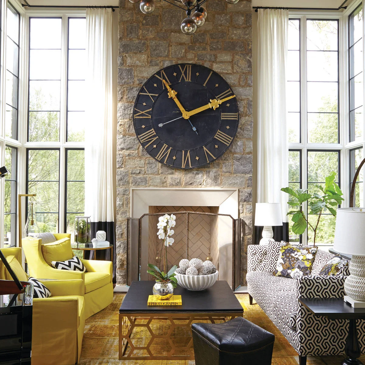 A tall stone fireplace wall with an oversized black clock, white curtains, yellow chairs and a black-and-white patterned sofa.